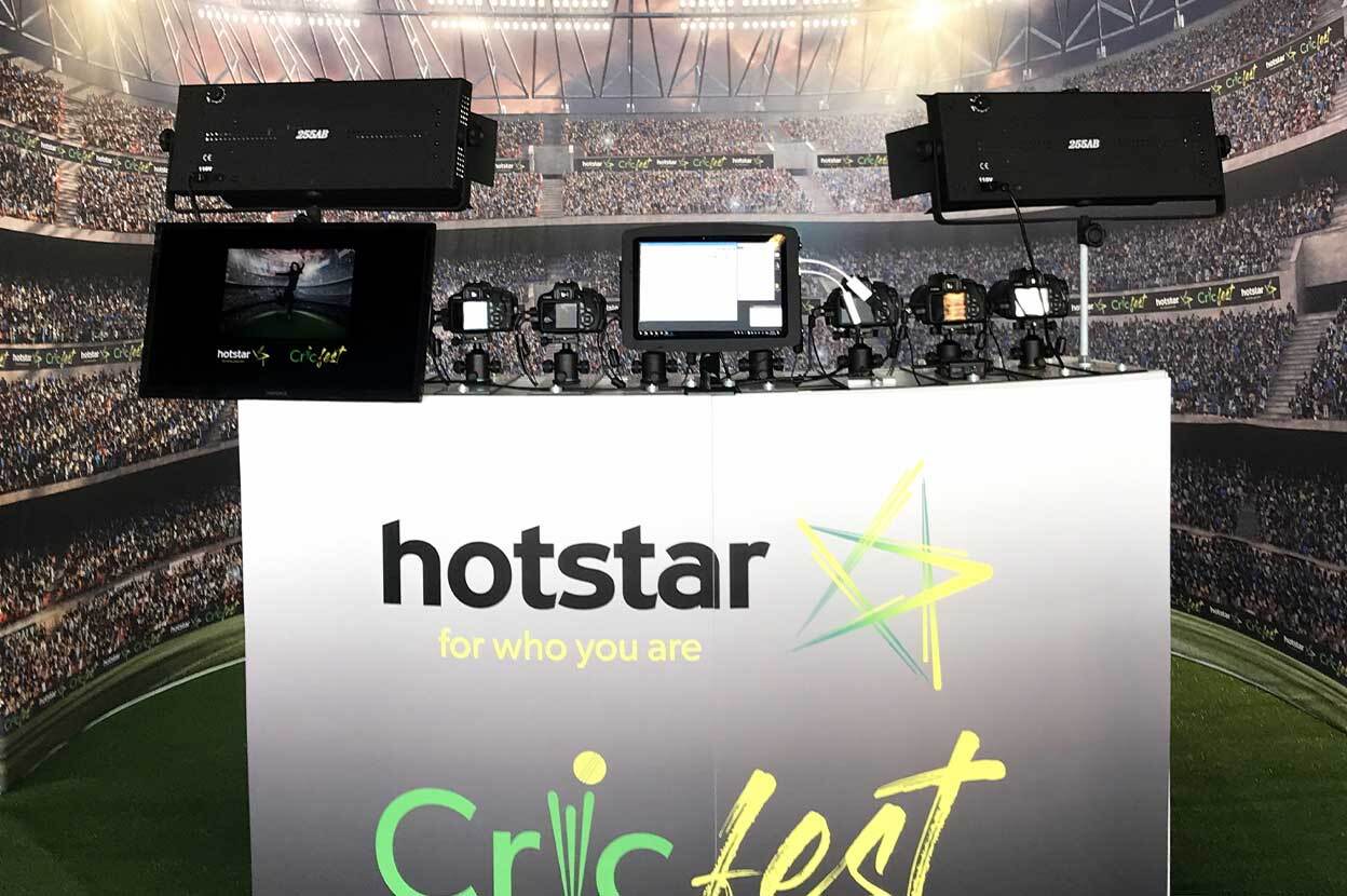 Backdrop of a full cricket stadium behind a podium with six cameras, video monitors, and professional lighting.