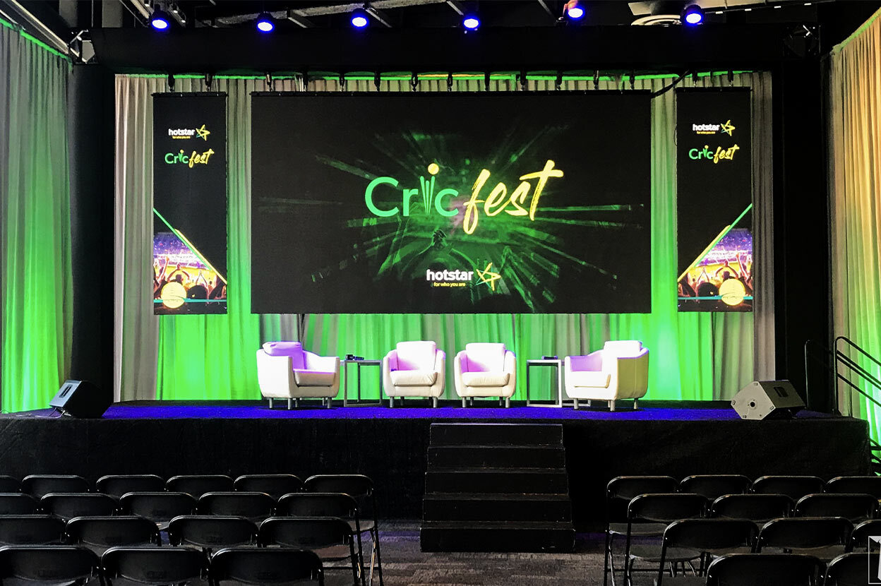 Stage with green accent lighting, Cricfest banners, and white armchairs.